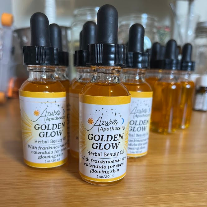 Three small glass dropper bottles labeled 'Azure's Apothecary Golden Glow Herbal Beauty Oil' arranged on a wooden surface, with more bottles blurred in the background.